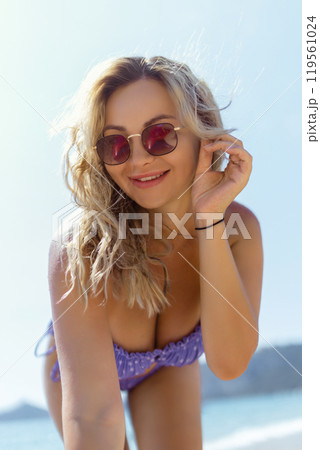 A beautiful girl in sunglasses on the beach looks at the camera, leaning towards it. Sexy blonde in a swimsuit on the seashore 119561024