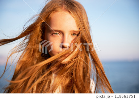 Portrait of a beautiful teenage girl with long flowing red hair against the background of the sea in summer at sunset. 119563751
