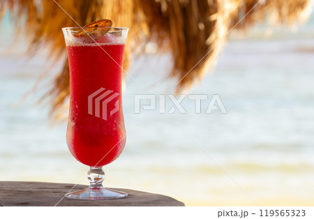 Cold red strawberry cocktail in a glass with a dry orange slice on the wooden table on the beach under the straw roof umbrella, the beach and the sea is on the background. 119565323