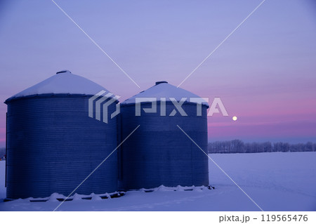 Picturesque winter sunrise in the grain farm in prairies. Granaries along the country road in purple and pink morning light. 119565476