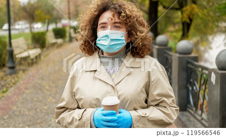 Pandemic walk. Outdoor quarantine. Sad depressed overweight woman with curly hair in medical mask strolling on empty autumn street. 119566546