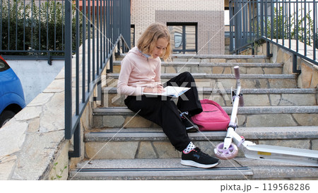 Schoolgirl studies her textbook on the steps of a residential building, her scooter resting nearby 119568286