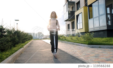 Cheerful schoolgirl rides her scooter down a sunlit street as she heads to school, enjoying the bright morning 119568288