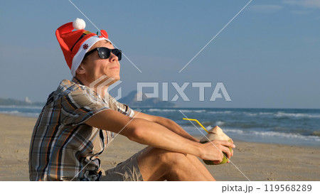 Man in a festive Christmas hat and sunglasses enjoys coconut water while lounging on a tropical beach during the winter holidays 119568289