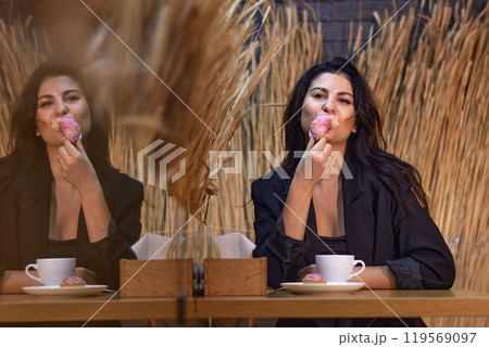 Woman eating, Pink donut, Coffee cafe. Woman indulges in pink-frosted donut while having coffee at cafe, with mirror reflection and rustic wheat decor creating cozy vibe. Woman eating, Pink donut, Coffee cafe. Woman indulges in pink-frosted donut while having coffee at cafe, with mirror reflection and rustic wheat decor creating cozy vibe. 119569097