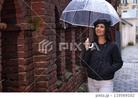 Stylish woman, clear umbrella, rainy day. Stylish woman walks down street with clear umbrella on rainy day, dressed in black quilted jacket white pants, smiles while strolling beside old brick wall Stylish woman, clear umbrella, rainy day. Stylish woman walks down street with clear umbrella on rainy day, dressed in black quilted jacket white pants, smiles while strolling beside old brick wall 119569102