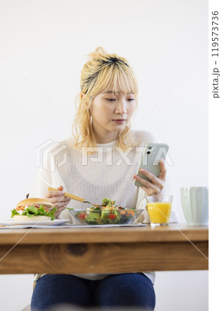 スマホを見ながら朝食を食べる女性 スマホを見ながら朝食を食べる女性 119573736