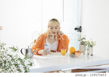 スマホを見ながら朝食をとる若い金髪の女性 スマホを見ながら朝食をとる若い金髪の女性 119573853