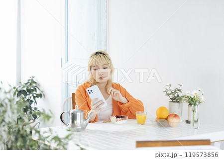 スマホを見ながら朝食をとる若い金髪の女性 スマホを見ながら朝食をとる若い金髪の女性 119573856