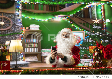 Santa Claus is online browsing gift ideas on his smartphone in a festive decorated cabin. Spreading the magic of Christmas at the North Pole, preparing for the journey, holiday traditions. Santa Claus is online browsing gift ideas on his smartphone in a festive decorated cabin. Spreading the magic of Christmas at the North Pole, preparing for the journey, holiday traditions. 119575345