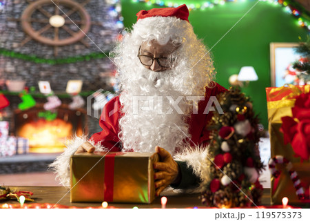 Santa Claus wraps a festive gift box by the fire in his North Pole workshop, surrounded by holiday lights and decorations. Father Christmas prepares gifts and surprises for winter celebration. Santa Claus wraps a festive gift box by the fire in his North Pole workshop, surrounded by holiday lights and decorations. Father Christmas prepares gifts and surprises for winter celebration. 119575373