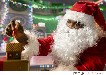 Father Christmas decorates homemade gingerbread house with frosting and beads, North Pole log cabin workshop. Santa Claus keeping the festive tradition of baking sweet treats with cinnamon. Father Christmas decorates homemade gingerbread house with frosting and beads, North Pole log cabin workshop. Santa Claus keeping the festive tradition of baking sweet treats with cinnamon. 119575377
