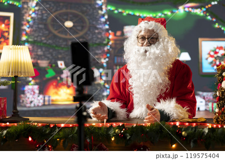 Father Christmas records a vlog talking to his online audience about the magic of Christmas. Santa Claus in a cozy festive cabin connects with subscribers through social media during wintertime. Father Christmas records a vlog talking to his online audience about the magic of Christmas. Santa Claus in a cozy festive cabin connects with subscribers through social media during wintertime. 119575404