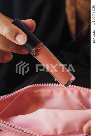 A fashionable hand is gracefully holding a lip gloss positioned above a vibrant pink makeup bag A fashionable hand is gracefully holding a lip gloss positioned above a vibrant pink makeup bag 119575836