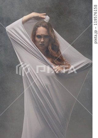 Female model draped in sheer fabric poses dramatically in a misty studio setting Female model draped in sheer fabric poses dramatically in a misty studio setting 119576158