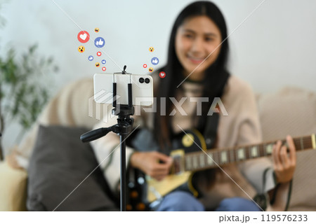 Young woman playing guitar during live streaming on smartphone at home Young woman playing guitar during live streaming on smartphone at home 119576233