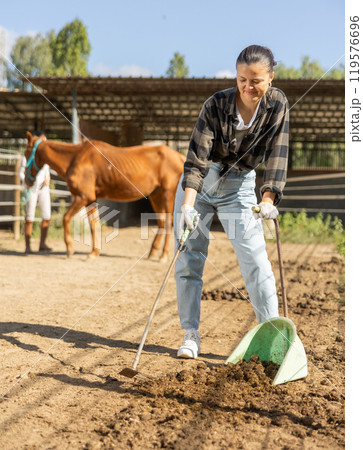 Smiling Asian girl worker scoops horse droppings into shovel and cleans up cattle 119576696