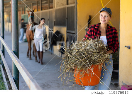 European young female worker walks go through corridor of stable, carrying basket with straw European young female worker walks go through corridor of stable, carrying basket with straw 119576915