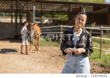 Asian female stable worker stands near wooden fence of paddock and watches horse. 119576931