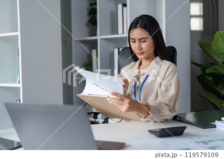 Business Professional Analyzing Documents in Modern Office Setting with Laptop and Calculator 119579109