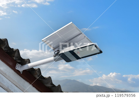 Mini solar cell panel and hd floodlight panal installed together under house eaves to store and use the power from the sunlight with street light and bulbs at night, sunset background, soft focus. Mini solar cell panel and hd floodlight panal installed together under house eaves to store and use the power from the sunlight with street light and bulbs at night, sunset background, soft focus. 119579356