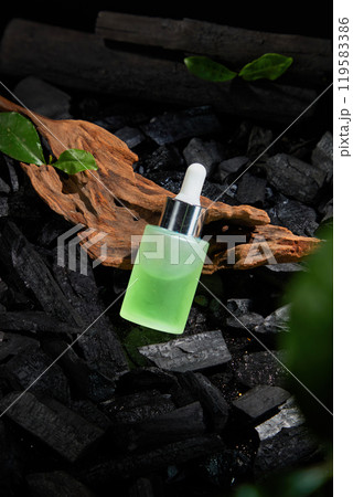 On a piece of weathered driftwood with some green leaves, and the charcoal fragments surrounding it. The image shows a mockup of a glass dropper bottle filled with a light green liquid. 119583386