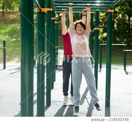 Elderly man and woman doing exercises on horizontal bars at outdoor sports ground 119583730