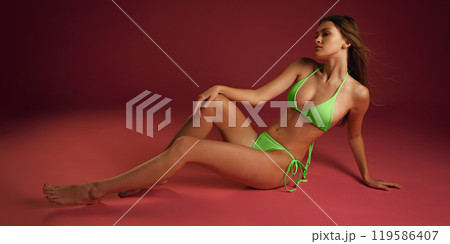Full-body shot of woman posing, lying confidently in vibrant green bikini against burgundy color background. Relax mood Full-body shot of woman posing, lying confidently in vibrant green bikini against burgundy color background. Relax mood 119586407