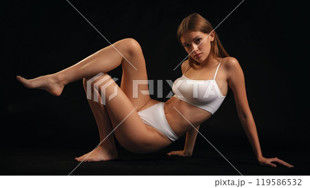 Slender young woman, dressed white lingerie posing, sitting , shows flexibility and balance against black studio background. 119586532