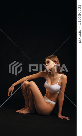 Slender young woman, dressed white lingerie posing, sitting , shows flexibility and balance against black studio background. 119586533