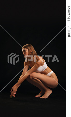 Young attractive slim woman posing, sitting in white lingerie against black studio background. 119586541