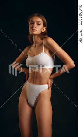 Young attractive woman posing, and looking at camera, in white lingerie holding hands on hips,against black studio background. Young attractive woman posing, and looking at camera, in white lingerie holding hands on hips,against black studio background. 119586649