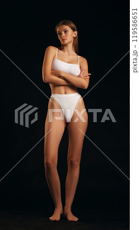 Full-body portrait of woman in white lingerie, crossing her arms, elegantly posing against black studio background. 119586651