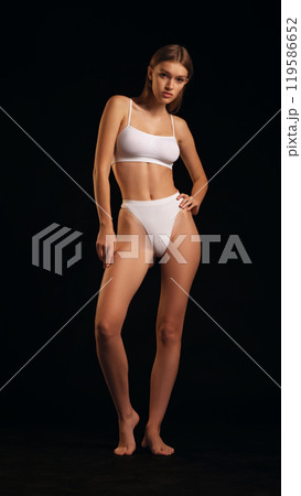 Full-body photo of beautiful, fit woman poses in white lingerie against black studio background. Woman with fit physique. 119586652