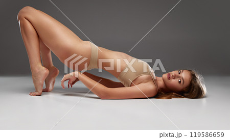 Young fit woman bridging her body upward, showing balance and strength, wearing neutral bikini against warm grey background. Young fit woman bridging her body upward, showing balance and strength, wearing neutral bikini against warm grey background. 119586659