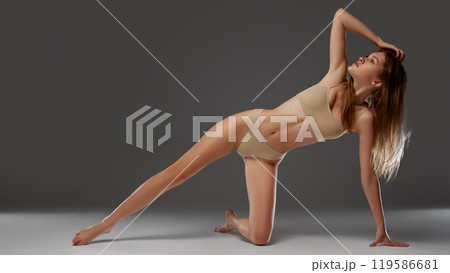 Fit, beautiful woman in side plank pose in nude bikini, striking dynamic yet elegant form against warm grey background. 119586681
