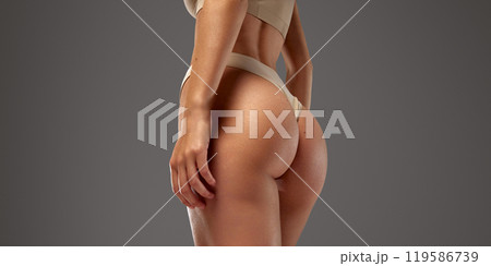 Young female model stands with her back turned, showcasing toned legs and back against warm grey background. 119586739