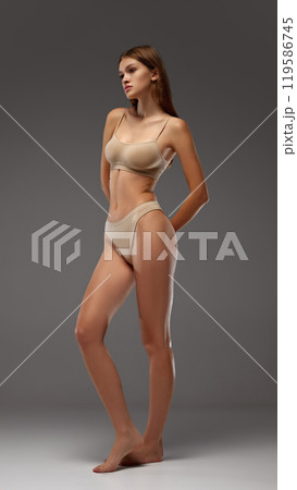 Young female model strikes thoughtful pose, showing off toned muscles against warm grey background. Full-body portrait. 119586745