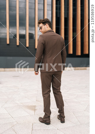 Back view of stylish young businessman in fashion brown suit and sunglasses confidently standing in city amidst modern architecture. Photogenic business man posing outdoors alone enjoying summer day. 119586919