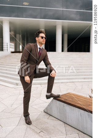 Vertical portrait of confident young businessman in stylish brown suit and sunglasses posing outdoors, exuding success and professionalism, city architecture complementing modern urban look. 119586920