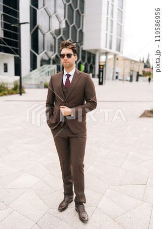 Vertical portrait of confident young businessman in stylish brown suit standing in front of modern office building. Handsome successful business man wearing sunglasses posing holding hand in pocket. Vertical portrait of confident young businessman in stylish brown suit standing in front of modern office building. Handsome successful business man wearing sunglasses posing holding hand in pocket. 119586956