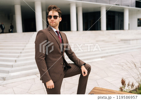 Portrait of confident successful businessman in brown suit and sunglasses standing in front of modern office building. Stylish businessman in elegant formal attire posing alone outdoors, looking away. Portrait of confident successful businessman in brown suit and sunglasses standing in front of modern office building. Stylish businessman in elegant formal attire posing alone outdoors, looking away. 119586957