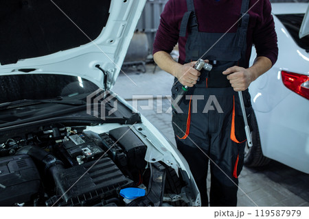 Car service. An auto mechanic is standing near the car with a tool in his hands. Vehicle technical inspection Car service. An auto mechanic is standing near the car with a tool in his hands. Vehicle technical inspection 119587979