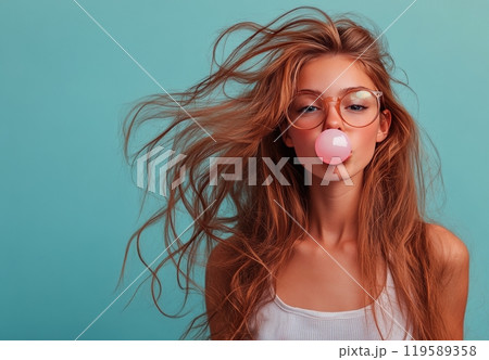 A young woman with long hair styled in waves enjoys blowing a bubble with pink gum 119589358