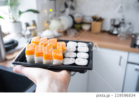 Delivery of sushi and rolls set is hold in hand in the interior modern kitchen of house Delivery of sushi and rolls set is hold in hand in the interior modern kitchen of house 119596834