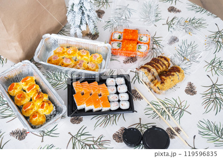 Delivery of sushi and rolls set top view on the table with a Christmas decor with a Christmas tree and a tablecloth with snowflakes, home cooking Delivery of sushi and rolls set top view on the table with a Christmas decor with a Christmas tree and a tablecloth with snowflakes, home cooking 119596835