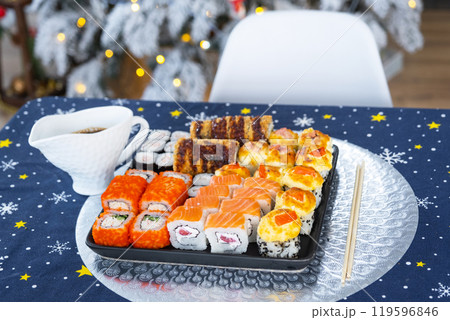 Delivery of sushi and rolls in food containers on the table in the festive interior of the kitchen of the house with a white Christmas tree for Christmas and New year. Food delivery for the holidays 119596846