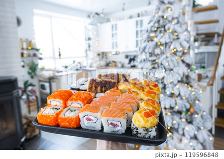Delivery of sushi and rolls set is hold in hand in the festive interior of house with a white Christmas tree for Christmas and New year. Food delivery for the holidays 119596848