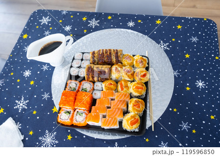 Delivery of sushi and rolls set top view on the table with a Christmas decor with a Christmas tree and a tablecloth with snowflakes, home cooking Delivery of sushi and rolls set top view on the table with a Christmas decor with a Christmas tree and a tablecloth with snowflakes, home cooking 119596850