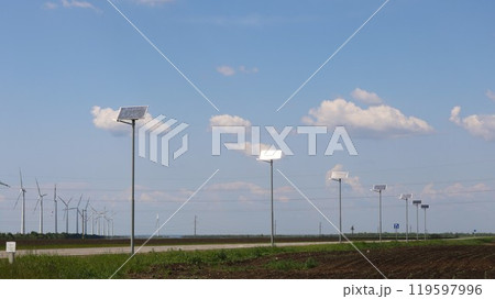 solar panels along highway with wind turbines in background, green energy of solar and wind power, renewable resources of planet earth, roadside panels and wind generators in fields 119597996